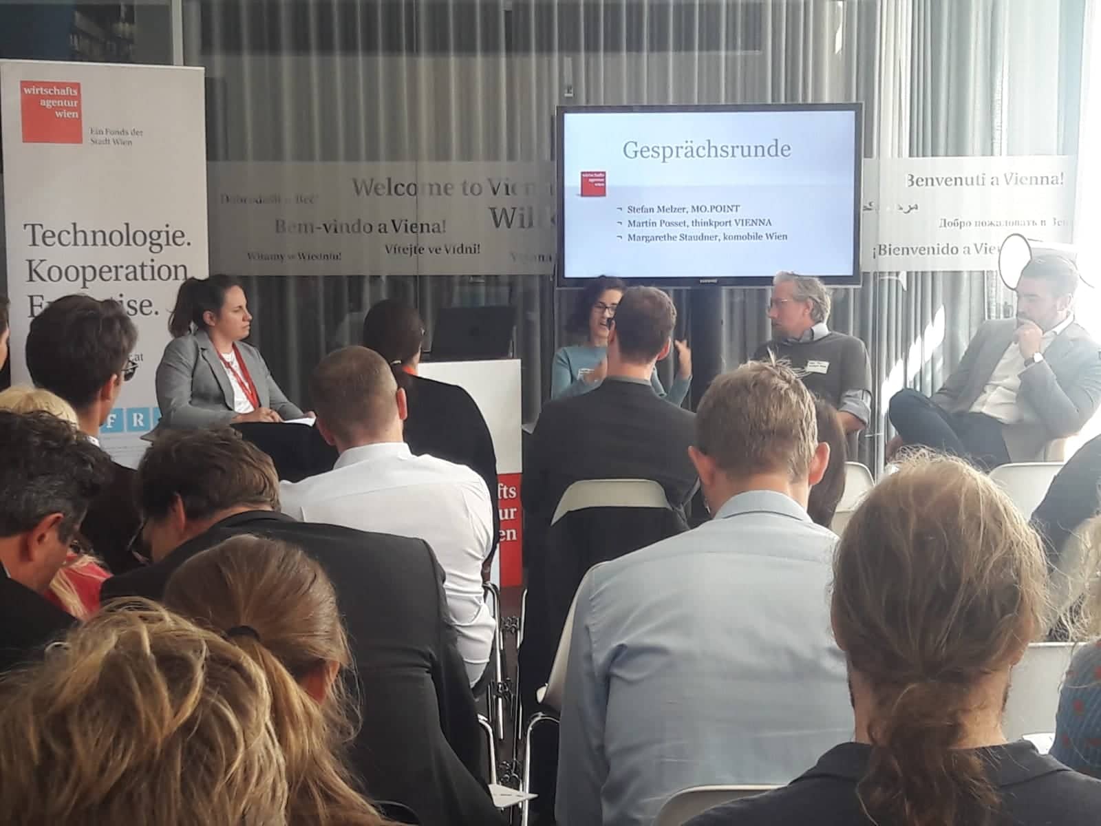 Die TeilnehmerInnen der Veranstaltungen sitzen in Reihen. Im vorderen Bereich des Bildes ist eine Podiumsdiksussionsrunde zu sehen.